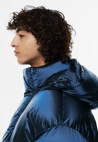 Podszewkowana niebieska kurtka puchowa o błyszczącej, pikowanej powierzchni oraz regulowanym kapturze. Posiada wysoki kołnierz i zaokrąglone szwy na ramionach.