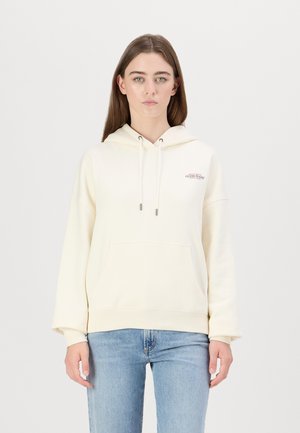 Cream-colored hoodie made of soft fabric with a front pocket, drawstring hood, and a small red logo on the chest. Paired with light denim jeans.
