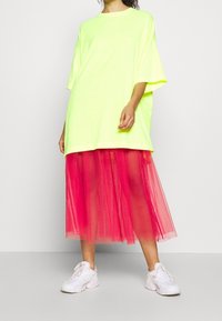 T-shirt oversize giallo neon con maniche corte, abbinato a una gonna in tulle rosa di lunghezza media. Il modello indossa sneakers bianche. Design semplice, colori vivaci.