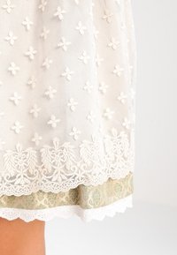 Jupe en dentelle blanche avec un délicat brodé floral, ourlet en dentelle festonnée et une couche inférieure texturée verte ornée de motifs élaborés.