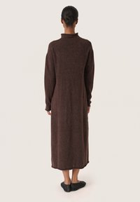 Langes, braunes Strickkleid mit hohem Kragen und langen Ärmeln, locker geschnitten mit seitlichen Schlitzen am Saum. Sichtbare schwarze Ballerinas.