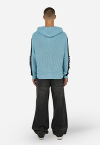 Šviesiai mėlyna raukšlėta ilgaaulė striukė su kapišonu, juodais šonų akcentais ir ilgomis rankovėmis, derinama su laisvomis tamsiai pilkomis kelnėmis.