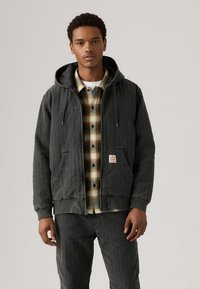 Gray hooded jacket with a quilted lining, front pockets, and ribbed cuffs. Underneath, a checkered flannel shirt in muted tones.