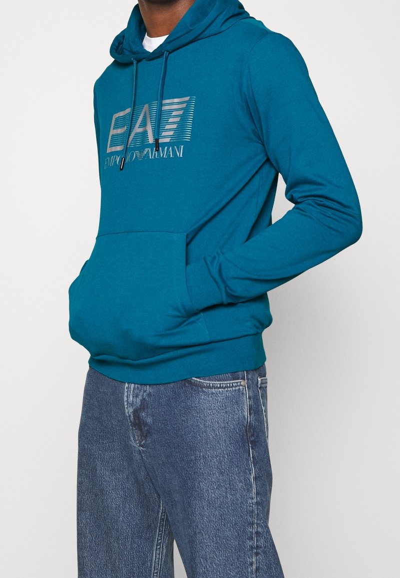 Homme portant un sweat à capuche teal Emporio Armani avec poche avant et jean bleu, debout les mains dans les poches devant un fond uni.