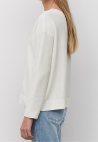 Weißer, gerippter Strickpullover mit langen Ärmeln, rundem Ausschnitt und niedrigem Saum. Kombiniert mit hellblauen Jeans. Glatte Textur, legerer Schnitt.