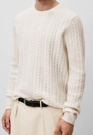 Mann trägt einen cremefarbenen, kabelgestrickten Pullover, in beige Hosen mit schwarzem Ledergürtel gesteckt, vor einem einfarbigen Hintergrund.