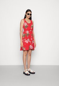 Vestido de sol vermelho com flores, cintura ajustada e saia rodada. Usado com óculos de sol pretos e sandálias brancas com tiras. Textura de tecido suave.