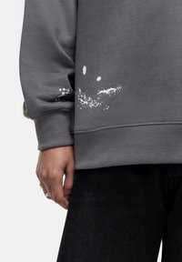 Grijze sweatshirt met witte spetters nabij de onderste zoom, met een ontspannen pasvorm en geribbelde manchetten. Gemaakt in combinatie met zwarte broek.