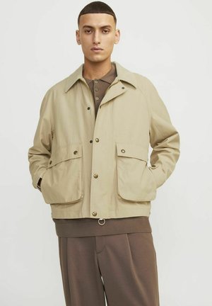 Beige kavaj med knäppning fram, två stora framfickor och en kontrasterande krage. Bärs över en brun ribbad tröja och byxor.