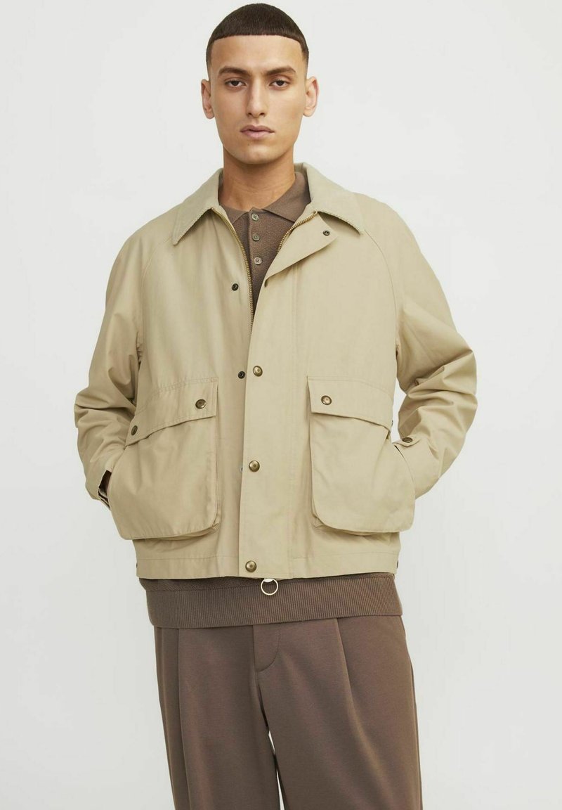 Beige jakke med knaplukning, to store frontlommer og en kontrastfarvet krave. Båret over en brun ribbestrikket sweater og bukser.