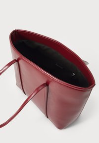 Sac fourre-tout en cuir rouge avec une texture lisse, ouverture en haut et deux poignées plates. L'intérieur est doublé de tissu noir avec une poche zippée.