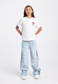 Wit katoenen T-shirt met een kleine rode afbeelding en zwarte omlijning op de linkerborst; lichtblauwe oversized spijkerbroek met contrasterende zijpanelen en witte sneakers.