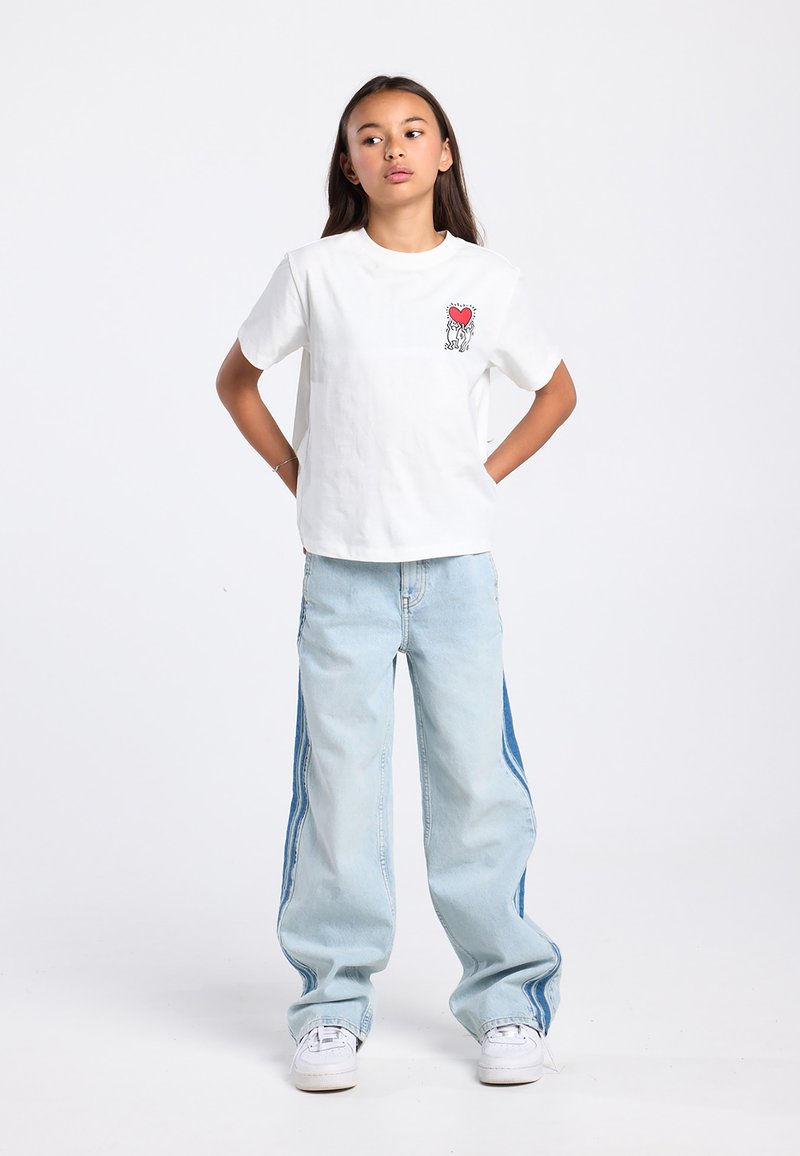 Wit katoenen T-shirt met een kleine rode afbeelding en zwarte omlijning op de linkerborst; lichtblauwe oversized spijkerbroek met contrasterende zijpanelen en witte sneakers.