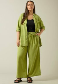 Groene linnen overhemd over een zwarte tanktop, gecombineerd met losse groene linnen broeken. Zwarte sandalen met een strikaccent. Gladde textuur.