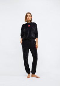Zwart velours loungewear set met een lange mouwen top met een rood logo label en jogger-stijl broek met een elastische tailleband en trekkoord.