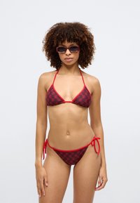 Rød bikiniset med trekantet topp og lavt liv, med mønster og røde bånd på sidene. Jevn stofftekstur.