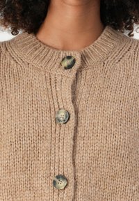 Beige chunky strikkegenser med knapper øverst, med runde marmorknapper med "Marc O'Polo" merking.