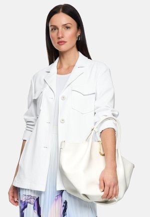 Frau mit langen dunklen Haaren, die eine weiße Jacke trägt und eine große weiße Handtasche hält, und einen plissierten Rock mit blau-lila Mustern.