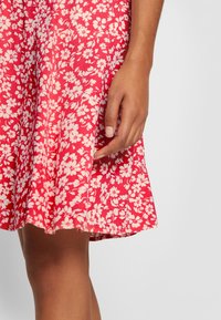 Röd blommig klänning med ett vitt blommönster, utsvängd fåll och lätt tyg. Hand delvis synlig, hållande klänningen.