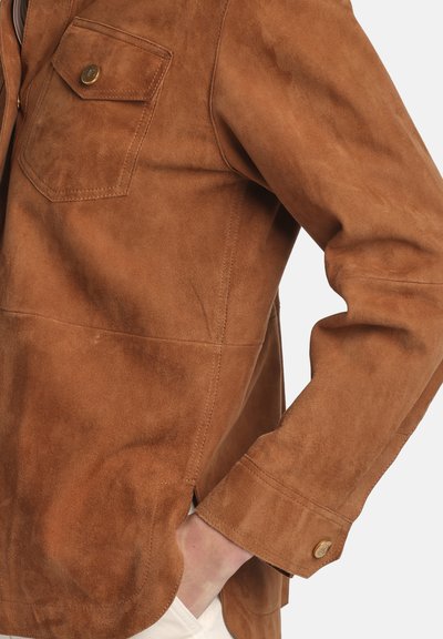 Braune Wildlederjacke mit zwei Brusttaschen, Knopfdetails und abgerundetem Saum. Weiche Textur und taillierte Passform sind hervorstechende Merkmale.