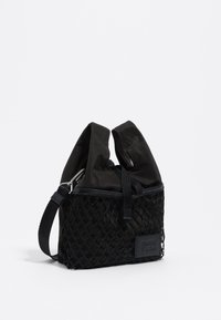 Bolsa negra con una base de malla texturizada y una parte superior lisa y brillante. Cuenta con un cierre de cordón y una correa ajustable. Parche con el logo en el lado.