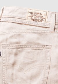 Svijetlo bež traperice s zakrivljenim dizajnom stražnjeg džepa, s smeđom kožnom oznakom marke ušivenom iznad pojasa.