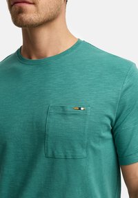 T-shirt a maniche corte color teal realizzato in morbido tessuto, con taschino sul petto decorato con dettagli a righe multicolori. Texture liscia e collo rotondo.