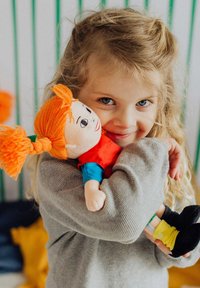Plüschpuppe mit orangefarbenem Garnhaar, trägt ein rotes Shirt und blaue Ärmel, gehalten von einem Kind in einem grauen Pullover, vor einem strukturierten Hintergrund.