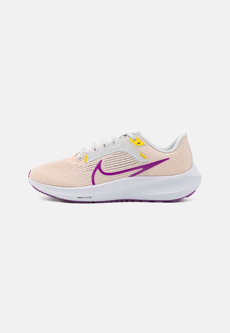 Chaussure de course beige clair avec semelle blanche, logo swoosh violet, semelle extérieure violette et détails jaunes aux œillets des lacets sur fond blanc.