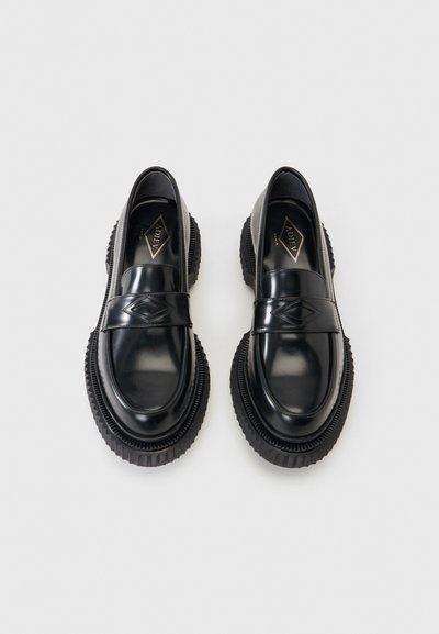 Ζευγάρι μαύρα γυαλιστερά δερμάτινα μοκασίνια με χονδρές ραβδωτές σόλες και διακοσμητικό σε σχήμα ρόμβου στο λουράκι, προβεβλημένα από πάνω σε λευκό φόντο.