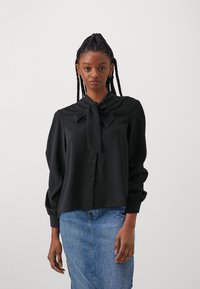 Svart bluse med sløyfe-kollare, knappelukking og lange pufferm; glatt tekstur og minimalistisk design kombinert med et denimskjørt.