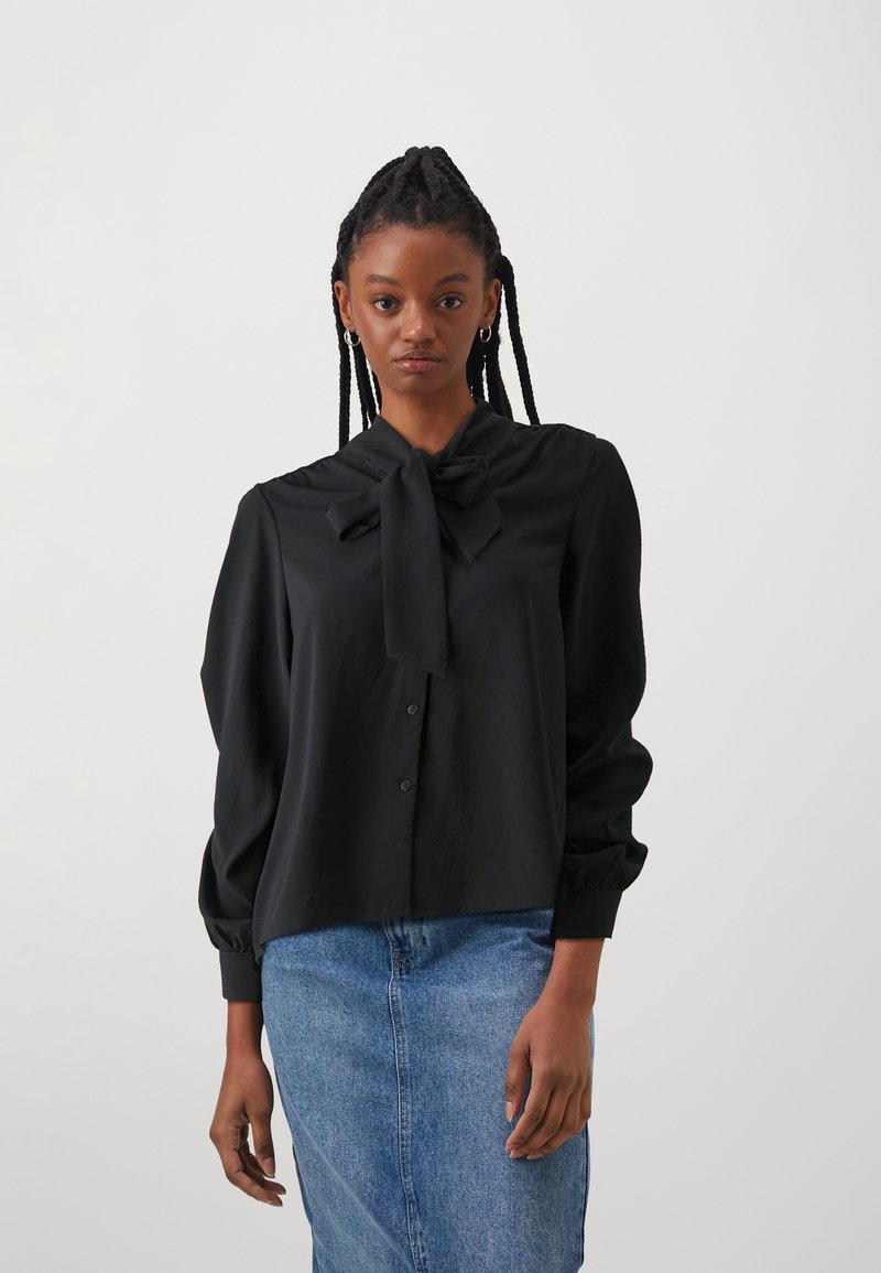 Svart bluse med sløyfe-kollare, knappelukking og lange pufferm; glatt tekstur og minimalistisk design kombinert med et denimskjørt.