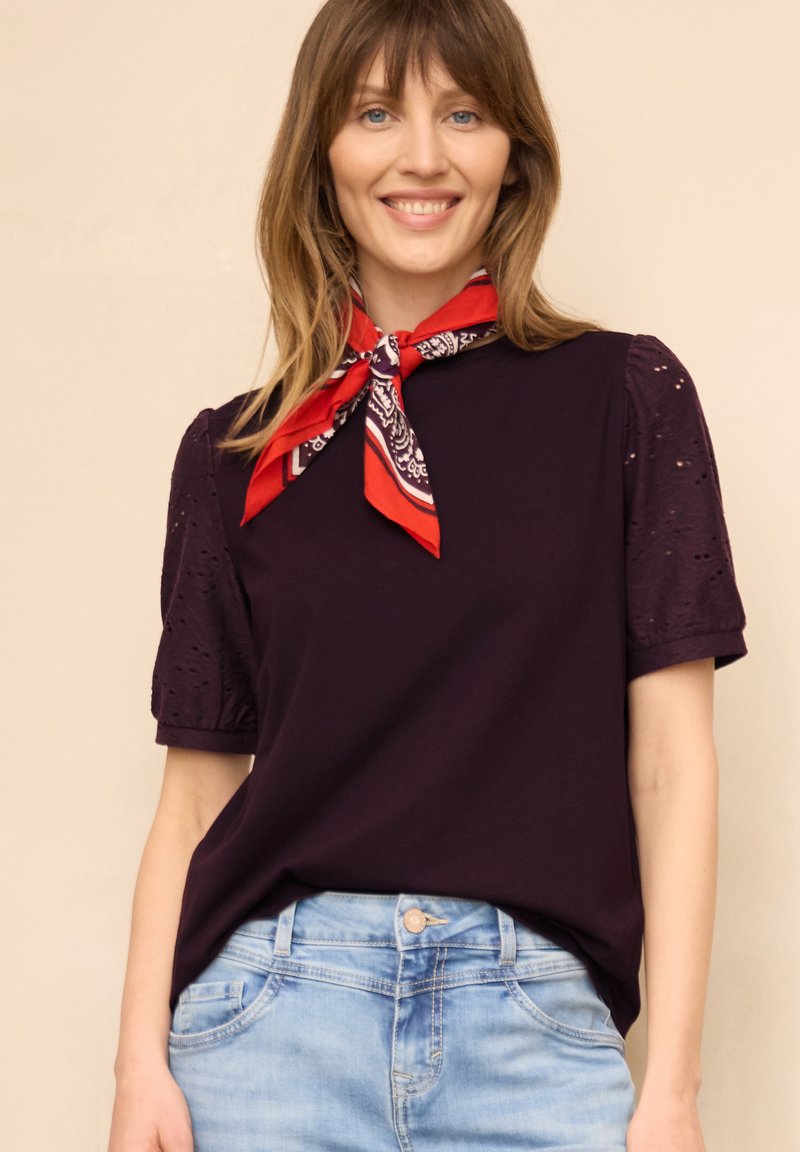 Femme portant un haut violet foncé avec manches bouffantes ajourées, un jean bleu clair et une écharpe rouge à motif nouée autour du cou, souriante.