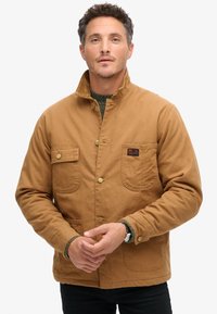 Chaqueta de algodón marrón con un cuello alto, dos bolsillos frontales y botones de latón. Tejido texturizado con un diseño casual.