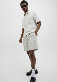 Chemise à manches courtes gris clair avec des boutons et une poche, associée à un short beige avec des poches latérales. Des chaussures noires et des lunettes de soleil complètent le look.