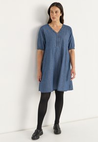 Denim jurk met een V-hals, korte puff mouwen en geraffineerde details. Kleur: middelblauw. Gecombineerd met zwarte tights en loafers.