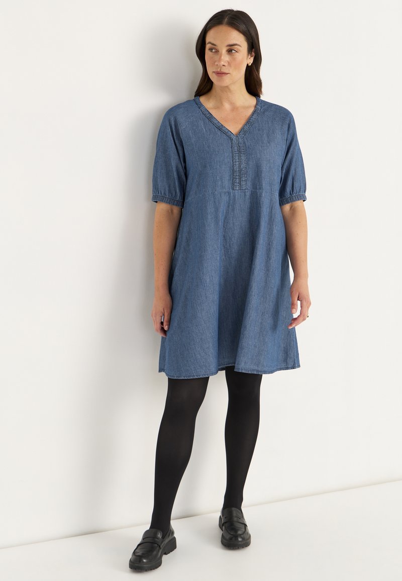 Denim jurk met een V-hals, korte puff mouwen en geraffineerde details. Kleur: middelblauw. Gecombineerd met zwarte tights en loafers.