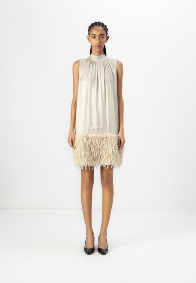 Robe sans manches de couleur crème avec un corsage plissé et une jupe texturée à franges beige. Le modèle porte des escarpins en vernis noir.