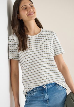 Frau mit langen braunen Haaren, die an einer Wand lehnt, trägt ein kurzärmliges, gestreiftes Strickoberteil und blaue Jeans und hält eine grüne Jacke in der Hand.