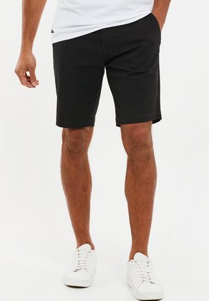 Homme portant un short noir jusqu'aux genoux, des baskets blanches et une chemise blanche, se tenant avec une main dans la poche devant un fond blanc.