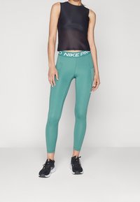 Musta hihaton toppi pyöreällä pääntiellä, yhdistettynä teal-värisiin leggings-housuihin, joissa on valkoinen Nike Pro -vyötärö ja sivutaskut. Mustat urheilukengät.