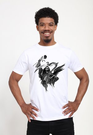 Glimlachende man die een wit shirt draagt met een zwart ontwerp van de Grim Reaper met een roos en zeis, staand met handen in de zij tegen een witte achtergrond.