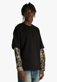 Maglietta nera oversize a maniche corte, indossata sopra una camicia a maniche lunghe con motivo e tonalità terrose. Il look è completato da jeans in denim.