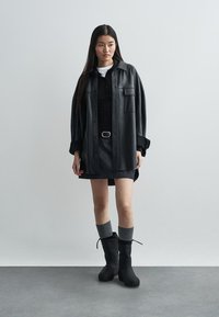 Cămașă jachetă supradimensionată din piele ecologică neagră, purtată peste un polo negru și o fustă închisă. Ciorapi gri până la genunchi asociați cu botine negre.