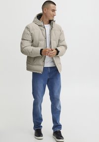 Light beige puffer jacket with a hood, featuring a quilted design, black accents, snap closures, worn over a white shirt and blue jeans.