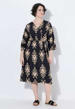 Frau mit kurzen dunklen Haaren, die ein schwarzes knielanges Kleid mit beigen Blumenmustern und schwarzen flachen Sandalen trägt, steht vor einem weißen Hintergrund.