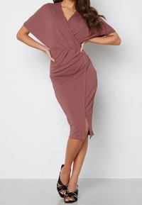 Robe ajustée mauve avec un design enveloppant et des manches courtes, présentant une fente sur le côté et des détails froncés. Portée avec des sandales à talons ouverts noires.