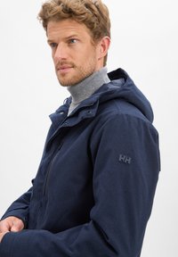 Dunkelblaue Kapuzenjacke aus strukturiertem Stoff mit einem Reißverschluss und Logoplatzierung auf dem linken Ärmel. Verfügt über einen hohen Kragen für zusätzliche Wärme.