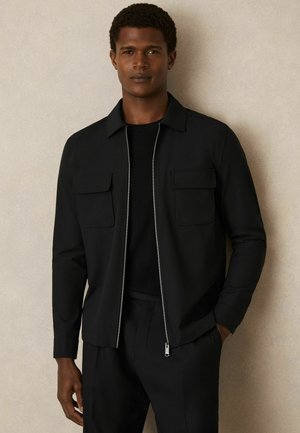 Jachetă neagră cu fermoar frontal, două buzunare pe piept și un guler structurat. Material neted cu un finisaj mat, asociat cu un tricou negru cu guler rotund.