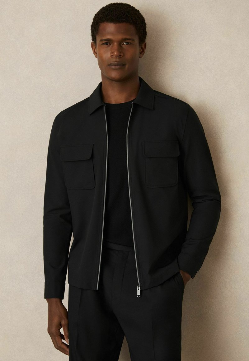 Zwarte jack met een rits aan de voorkant, twee borstzakken en een gestructureerde kraag. Gladde stof met een matte afwerking in combinatie met een zwart T-shirt met ronde hals.